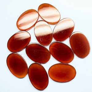 Peut inclure: Un ensemble de pierres précieuses ovales, translucides, de couleur ambre. Les pierres polies sont disposées sur une surface blanche, mettant en valeur leur finition brillante et leurs variations de couleur, allant de l'orange clair au brun rougeâtre foncé.