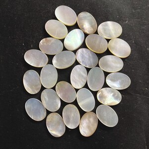 May include: A collection of oval-shaped, iridescent mother-of-pearl cabochons. The polished gemstones display a range of colors, including white, cream, and hints of blue and green. The cabochons are arranged on a dark surface.