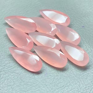 May include: A collection of nine faceted, teardrop-shaped gemstones in a soft pink hue. The gems are arranged on a light gray surface, each catching the light with multiple facets. The overall effect is one of delicate beauty and refined craftsmanship.