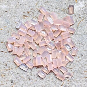 Könnte beinhalten: Eine Ansammlung rechteckiger, hellrosa Edelsteine auf einer strukturierten Oberfläche. Die Edelsteine haben ein facettiertes Aussehen, fangen das Licht ein und zeigen einen subtilen Schimmer. Die Anordnung deutet auf Rohmaterialien für Schmuck oder Bastelarbeiten hin.