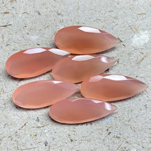 May include: Five pear-shaped, faceted gemstones in a soft peach-pink hue. Each stone features a smooth, polished surface with multiple geometric facets that reflect light, creating a translucent appearance across the teardrop-shaped silhouettes.