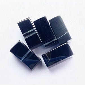 May include: Five rectangular black gemstones with polished surfaces and subtle white veining. The stones are arranged on a white surface, showcasing their glossy texture and natural patterns. These could be used for jewelry making.