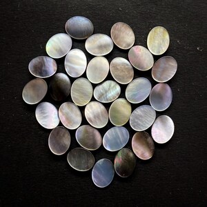 May include: A collection of oval-shaped mother-of-pearl cabochons. The iridescent stones display a range of colors, including white, gray, and hints of blue and green. The cabochons are arranged closely together, creating a visually appealing display.