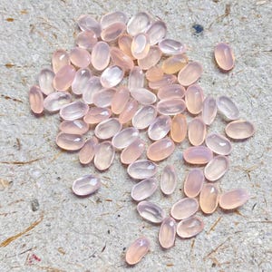 May include: A collection of numerous small, oval-shaped, translucent light pink gemstones. Each stone features a faceted surface that reflects light, creating a shimmering appearance across the group of loose, polished cabochons.