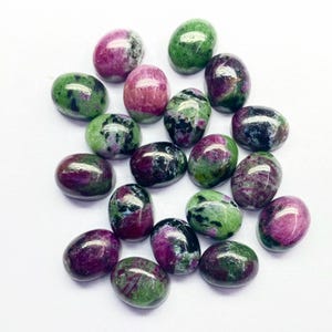 May include: A collection of polished oval gemstones in shades of deep red, green, and black. The stones have a smooth, glossy finish and vary in size. The gemstones are scattered on a white surface.