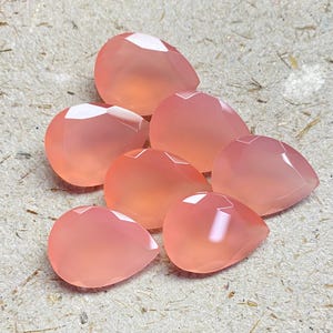 May include: A collection of six pear-shaped, faceted gemstones in a soft, translucent peach-pink hue. Each stone features a smooth, polished surface with geometric facets that catch the light, creating a subtle glow across the rounded edges.