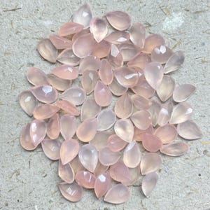 May include: A collection of teardrop-shaped, faceted gemstones in varying shades of pink. The gemstones are scattered across a neutral surface, with light reflecting off their cut surfaces. The overall effect is delicate and elegant.