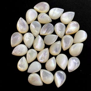 Può includere: Una collezione di cabochon a forma di goccia in madreperla iridescente. Le pietre preziose, lucidate e bianche, hanno una superficie liscia e riflettente con sottili variazioni di colore. I cabochon sono disposti su uno sfondo scuro.