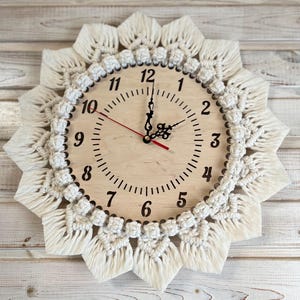 May include: A white macrame clock with a wooden face and black numbers. The clock is hanging on a white wooden wall.
