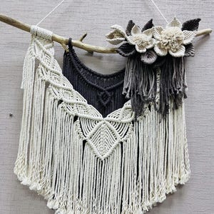 Peut inclure: Une suspension murale en macramé avec un motif géométrique noir et blanc et une frange blanche. La suspension est fixée à une branche avec trois fleurs de coton blanches.