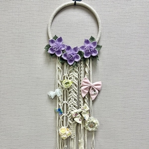 Puede incluir: Un soporte de lazo para el pelo de macramé con un marco circular color crema, adornado con tres acentos de flores moradas y hojas verdes. El soporte presenta hebras colgantes con varios lazos y adornos florales en colores pastel.