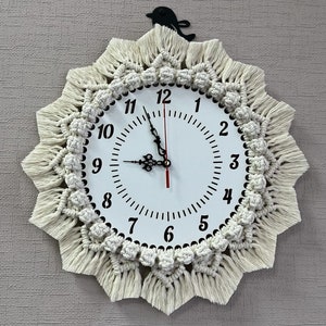 May include: A white clock with black numbers and hands, surrounded by a cream-colored macrame frame with fringe. A black bird silhouette is attached to the top of the clock.