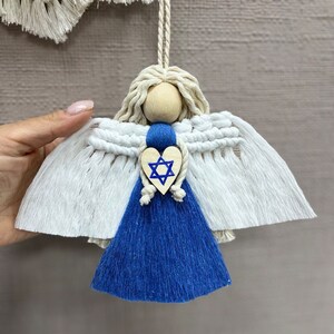 Könnte beinhalten: Weißer Makramee-Engel-Anhänger mit blauem Quasten und einem Holzhäuschen mit einem blauen Davidstern.