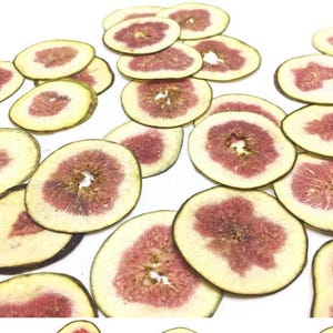 May include: A collection of thinly sliced dried fig rounds scattered across the frame. Each circular slice features a pale yellow-green outer skin and a textured, deep reddish-pink interior pulp with visible seeds radiating from the center.