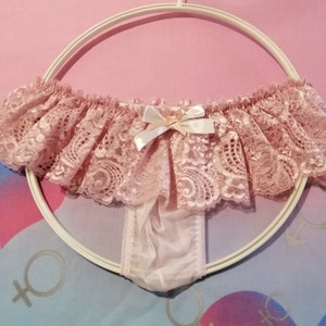 Pu&ograve; includere: Intimo in pizzo rosa con un fiocco sul davanti. L'intimo &egrave; appeso a un'appendiabiti bianca.