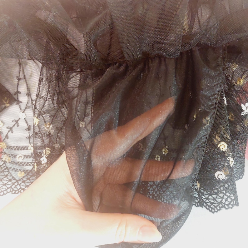 May include: Close-up of black tulle fabric with delicate gold sequins and lace trim. The sheer material is gathered, creating a layered effect. The lace features a floral pattern, adding a touch of elegance to the design.