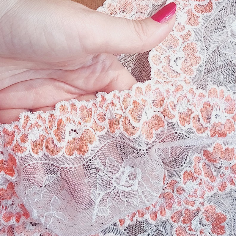 Puede incluir: Tela de encaje blanca con un delicado patr&oacute;n floral de color melocot&oacute;n. El encaje tiene un borde rizado y es suave y transparente.