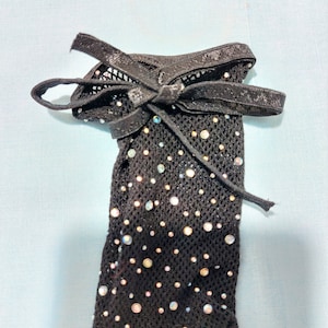 Puede incluir: Ligadura de encaje negra con un lazo de cinta negro y diamantes de imitaci&oacute;n iridiscentes.