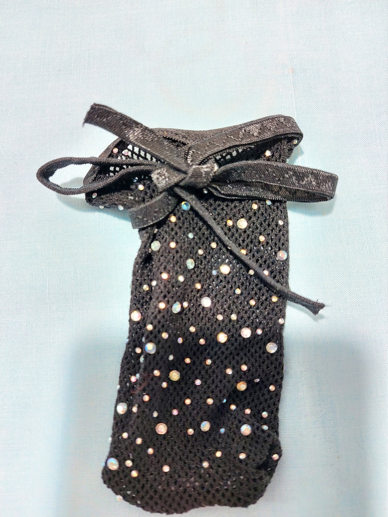 Puede incluir: Ligadura de encaje negra con un lazo de cinta negro y diamantes de imitaci&oacute;n iridiscentes.