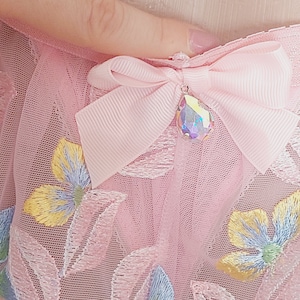 May include: Pink tulle fabric with embroidered floral designs and a pink bow with a clear iridescent crystal pendant.