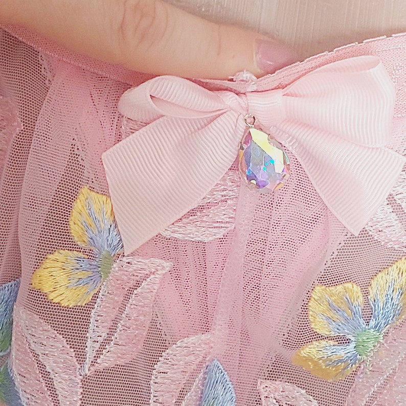 May include: Pink tulle fabric with embroidered floral designs and a pink bow with a clear iridescent crystal pendant.