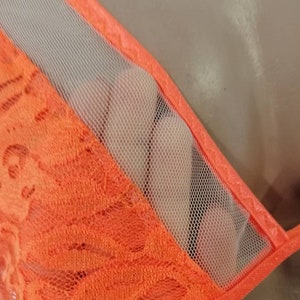 May include: Orange lace fabric with a sheer mesh panel. The lace has a delicate floral pattern.