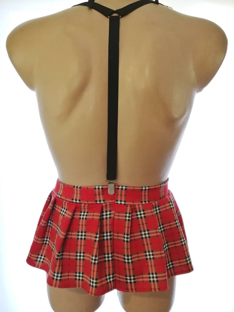 Puede incluir: Una minifalda roja y negra a cuadros con una tira de tirantes negra. La falda es plisada y tiene una cintura ajustada.