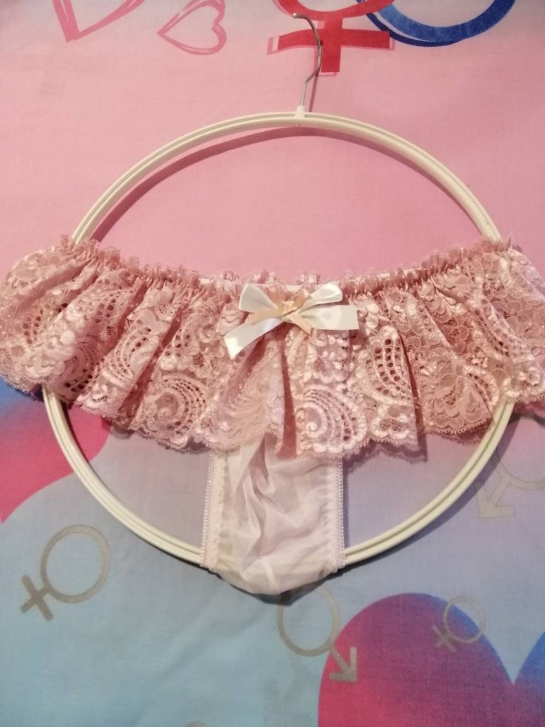 Pu&ograve; includere: Intimo in pizzo rosa con un fiocco sul davanti. L'intimo &egrave; appeso a un'appendiabiti bianca.