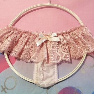 Pu&ograve; includere: Intimo in pizzo rosa con un fiocco bianco e un pannello trasparente sul retro. L'intimo &egrave; appeso a un appendiabiti bianco.