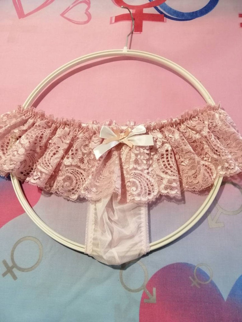 Pu&ograve; includere: Intimo in pizzo rosa con un fiocco bianco e un pannello trasparente sul retro. L'intimo &egrave; appeso a un appendiabiti bianco.