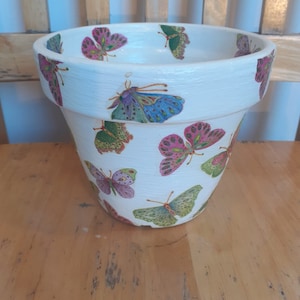 Könnte beinhalten: Weißer Keramik-Blumentopf mit Schmetterlingsdesign. Der Topf ist mit bunten Schmetterlingen in den Farben Rosa, Grün, Blau und Lila verziert.