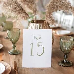 Puede incluir: Una tarjeta blanca con el texto "Tisch 15" escrito en caligrafía verde. La tarjeta está sobre una mesa con un jarrón de hierba de la pampa seca en el fondo.