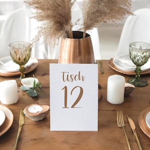 Puede incluir: Una tarjeta blanca con el texto "Tisch 12" sobre una mesa de madera con velas, platos y vasos. La mesa está preparada para una boda u otro evento especial.