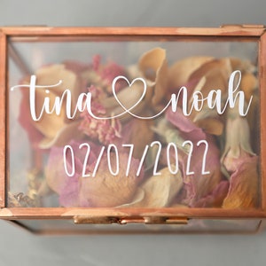 Op de afbeelding: Een transparante glazen doos met een koperen frame en een scharnierend deksel. De doos is gevuld met gedroogde rozenblaadjes en heeft een witte inscriptie met de tekst "tina & noah 02/07/2022".