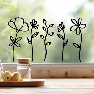 Op de afbeelding: Een zwarte vinyl muursticker met vijf verschillende florale lijntekeningen, waaronder een grote bloem, een madeliefachtige bloem, een stengel met knoppen, een kleine bloesem en een eenvoudige bloemvorm op een transparant glazen raam.
