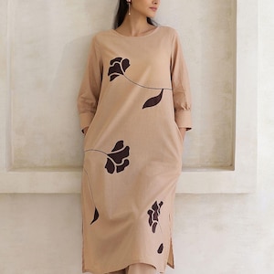 May include: A beige, long-sleeved dress and matching pants set. The dress features a round neckline and is adorned with dark brown floral designs. The outfit is made of a soft, flowing fabric, perfect for a casual, yet stylish look.