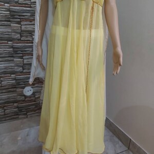 Pu&ograve; includere: Un abito giallo chiaro e fluido con un rivestimento trasparente e finiture con perline dorate. L'abito presenta un design senza maniche ed &egrave; abbinato a pantaloni palazzo bianchi. Uno scialle bianco e trasparente &egrave; drappeggiato sulle spalle.