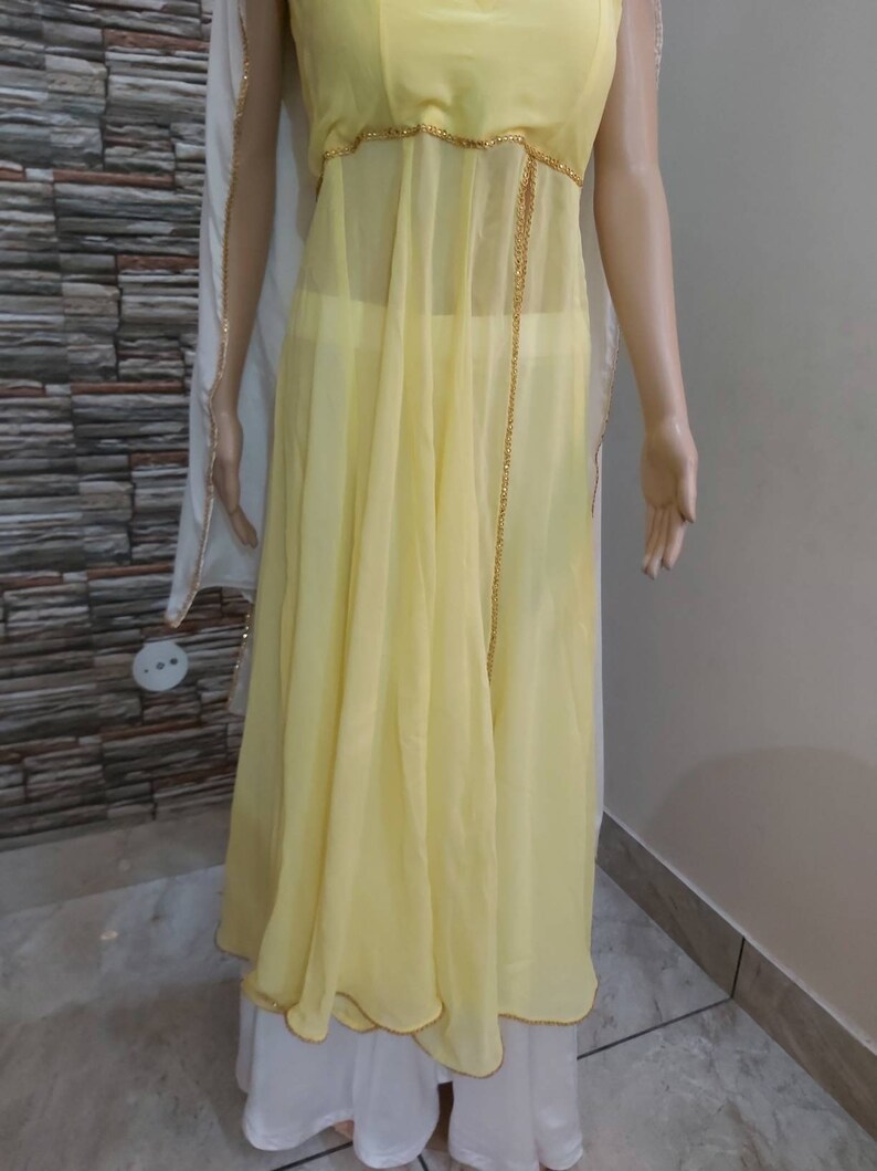 Pu&ograve; includere: Un abito giallo chiaro e fluido con un rivestimento trasparente e finiture con perline dorate. L'abito presenta un design senza maniche ed &egrave; abbinato a pantaloni palazzo bianchi. Uno scialle bianco e trasparente &egrave; drappeggiato sulle spalle.