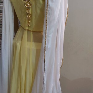 Pu&ograve; includere: Un vestito giallo con uno scialle bianco. L'abito ha uno scollo tondo e una gonna fluida. Lo scialle bianco &egrave; rifinito con perline dorate. Un elemento decorativo dorato pende dalla schiena del vestito.