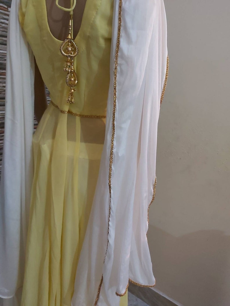Pu&ograve; includere: Un vestito giallo con uno scialle bianco. L'abito ha uno scollo tondo e una gonna fluida. Lo scialle bianco &egrave; rifinito con perline dorate. Un elemento decorativo dorato pende dalla schiena del vestito.