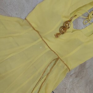 Pu&ograve; includere: Un abito giallo pallido con una cintura a catena color oro e abbellimenti decorativi. L'abito ha un tessuto fluido e leggero e uno scollo semplice. L'abbellimento dorato &egrave; dettagliato con piccoli accenti rotondi.