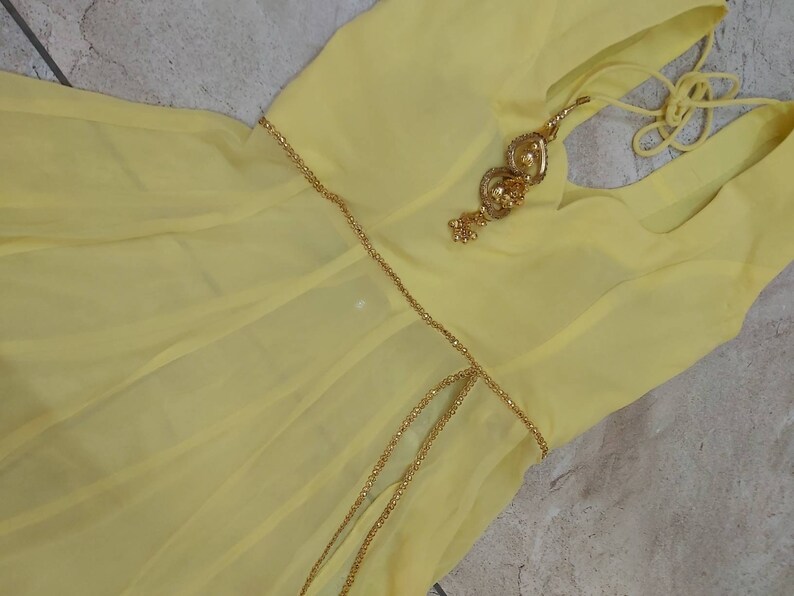 Pu&ograve; includere: Un abito giallo pallido con una cintura a catena color oro e abbellimenti decorativi. L'abito ha un tessuto fluido e leggero e uno scollo semplice. L'abbellimento dorato &egrave; dettagliato con piccoli accenti rotondi.