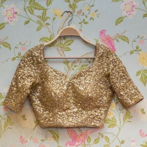 May include: Gold sequin blouse with a sweetheart neckline and short sleeves. The top is covered in small, shiny sequins, creating a shimmering effect. The blouse is hanging on a wooden hanger against a floral wallpaper background.
