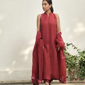 May include: A sleeveless, burgundy-colored dress with a high neckline and a row of buttons down the front. The dress is paired with matching pants and a sheer scarf. The outfit is completed with sandals. The ensemble is set against a white wall and greenery.