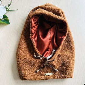 May include: A brown, hooded winter accessory with a soft, textured exterior and a satin lining. The hood features a drawstring with leopard print accents and a small gold-colored tag.
