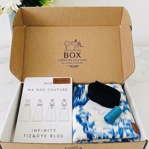 Puede incluir: Una caja de cartón que contiene un kit de costura con tela blanca y azul tie-dye, una banda elástica negra, un carrete de hilo azul y una tarjeta de instrucciones MA BOX COUTURE INFINITY TIE&DYE BLEU con ilustraciones de estilos de vestidos.