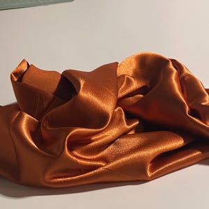 May include: A close-up shot of a crumpled piece of shiny, copper-coloured satin fabric. The fabric has a smooth texture and reflects light, creating highlights and shadows. The folds and curves of the fabric add depth and visual interest.