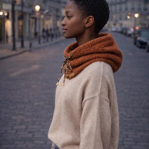 May include: A person wearing a tan sweater and blue jeans, with a rust-colored fleece cowl neck warmer. The cowl has a drawstring with a leopard print pattern and gold-colored accents. The background is a city street.