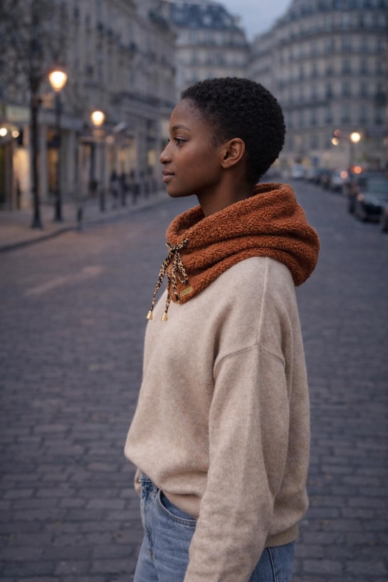 May include: A person wearing a tan sweater and blue jeans, with a rust-colored fleece cowl neck warmer. The cowl has a drawstring with a leopard print pattern and gold-colored accents. The background is a city street.