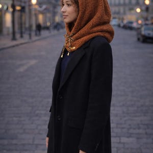May include: A woman wearing a brown hooded scarf with a decorative tie, paired with a black coat. The scarf is made of a textured material. The background shows a city street with streetlights.